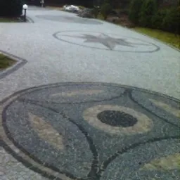 Ułożona kostka brukowa z dekoracyjnymi wzorami (gwiazda, okrąg) na posesji z budynkiem w tle. Widok z góry, oświetlenie dzienne.