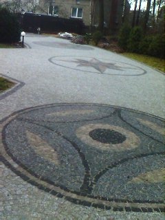 Ułożona kostka brukowa z dekoracyjnymi wzorami (gwiazda, okrąg) na posesji z budynkiem w tle. Widok z góry, oświetlenie dzienne.