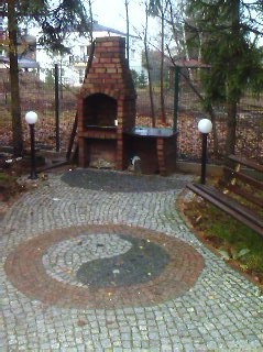 Ogród z brukowanym chodnikiem w kształcie Yin Yang, ceglany grill z kominem, lampy kuliste i ławka na tle drzew. Całość w jesiennej scenerii.