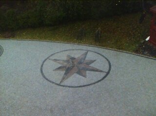 Szara kostka brukowa z dekoracyjnym wzorem gwiazdy w okręgu, otoczona zielenią. Widok z góry, lekko rozmyty obraz. Ciekawy detal brukowy.