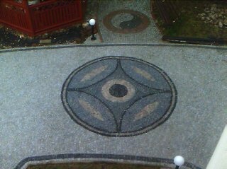 Dekoracyjny wzór z kamieni na podjeździe, okrągły motyw w centrum, szara kostka brukowa, widok z góry, oświetlenie zewnętrzne.