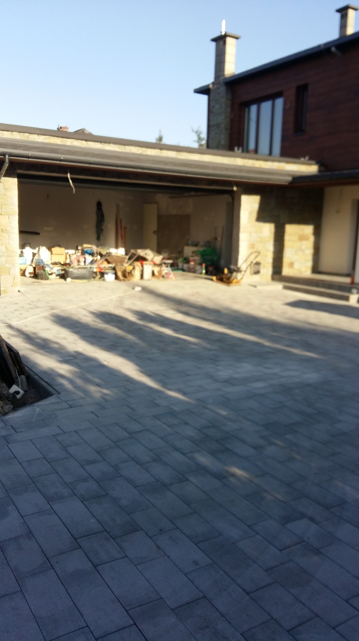 Ułożona kostka brukowa na podjeździe, z cieniem drzew i widokiem na otwarty garaż z narzędziami w tle, w słoneczny dzień.