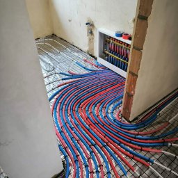OliArt Instalacje Oleksandr Shram - Rozłożona instalacja ogrzewania podłogowego z czerwono-niebieskimi rurami na folii aluminiowej, widoczny rozdzielacz w białej obudowie i surowe ściany w tle.