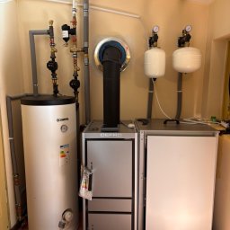 OliArt Instalacje Oleksandr Shram - Nowoczesna kotłownia z piecem DEFRO i pionowym podgrzewaczem KOSPEL, widoczne rury, zawory, pompy i dwa naczynia wzbiorcze. Kompletny system grzewczy na tle żółtych ścian i terakoty.