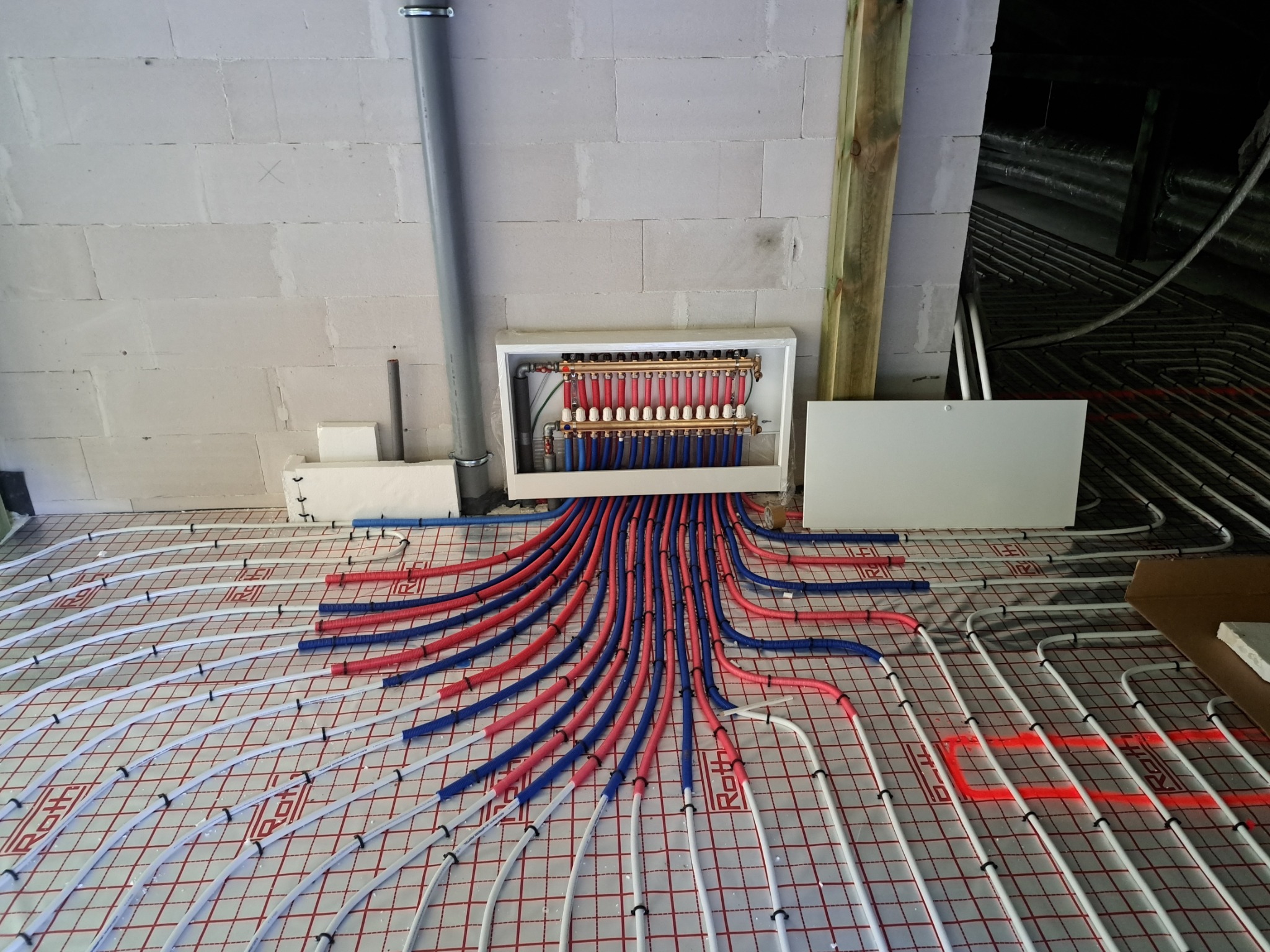 Rozdzielacz ogrzewania podłogowego z czerwono-niebieskimi rurami na białej macie z siatką, zamocowane czarnymi klipsami. Ściana z betonu komórkowego w tle.