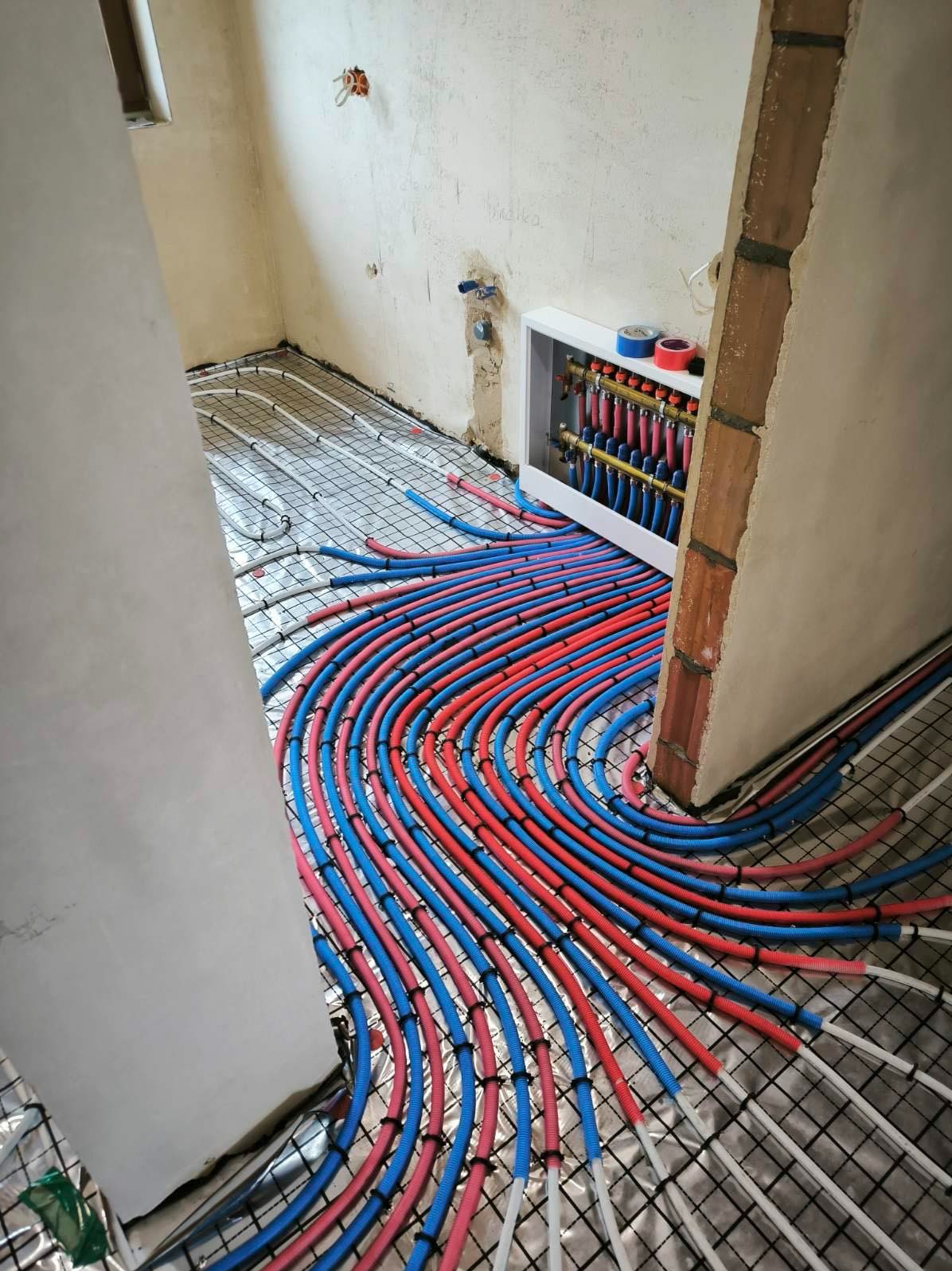 Rozłożona instalacja ogrzewania podłogowego z czerwono-niebieskimi rurami na folii aluminiowej, widoczny rozdzielacz w białej obudowie i surowe ściany w tle.