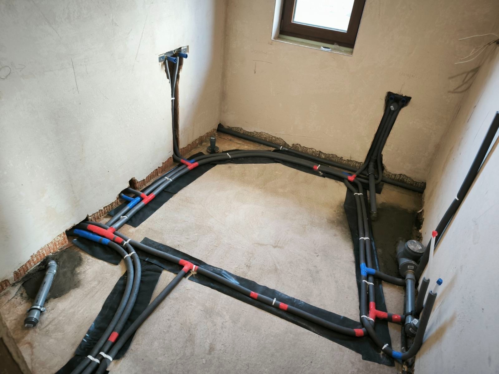 Szare rury instalacji grzewczej/hydraulicznej ułożone na czarnej folii w surowym pomieszczeniu. Widoczne połączenia w kolorze czerwonym i niebieskim.