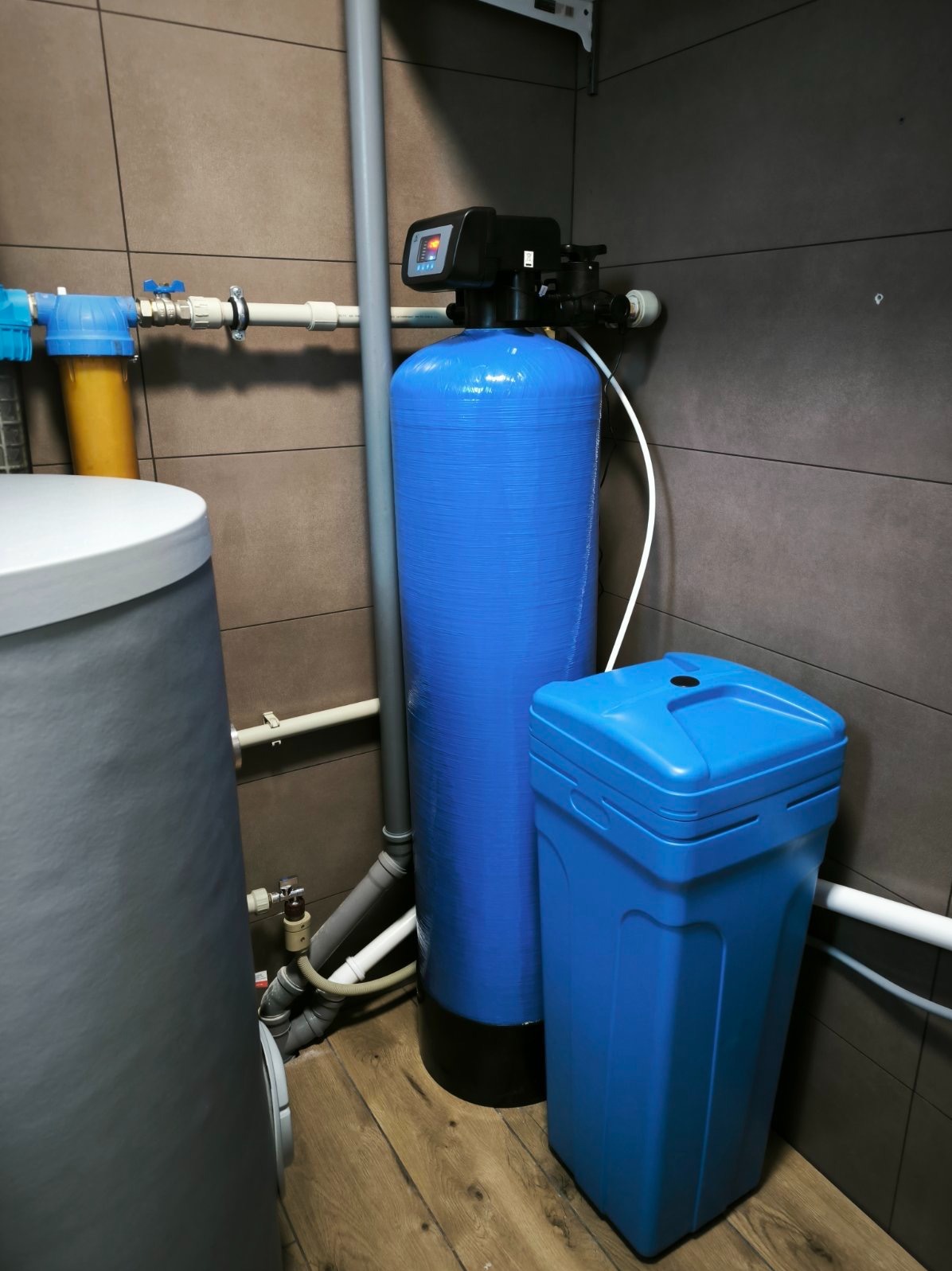 Niebieski zmiękczacz wody z filtrem wstępnym i zbiornikiem na sól, zainstalowany w pomieszczeniu z brązowymi płytkami i drewnianą podłogą.