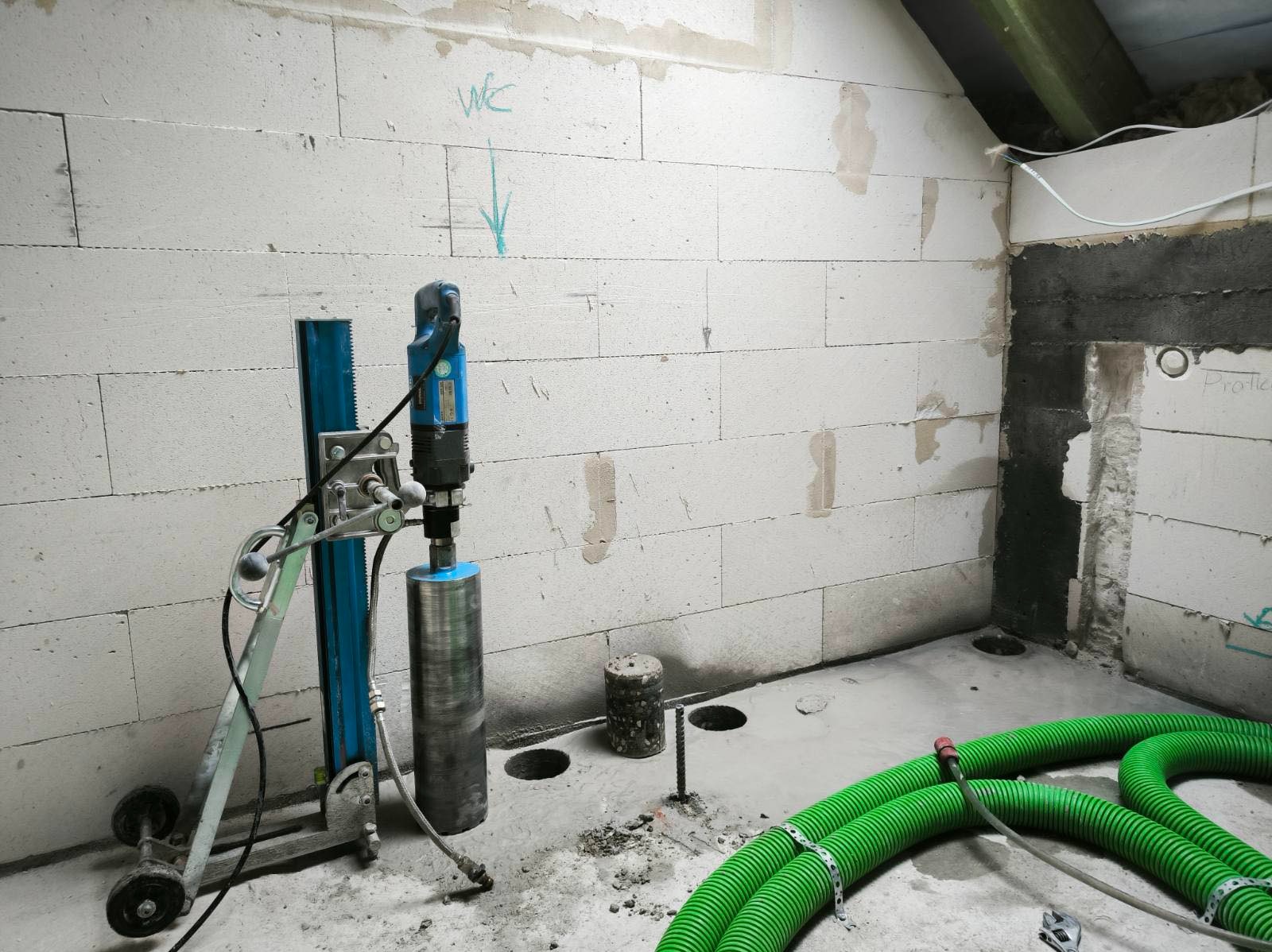 Wiertnica diamentowa do betonu, otwory w posadzce, zielone rury karbowane. Przygotowanie do instalacji w pomieszczeniu z białych bloczków.