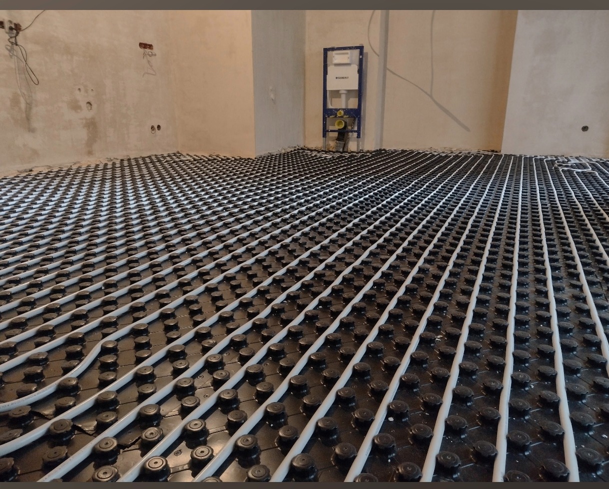 Ułożona instalacja ogrzewania podłogowego w systemie mokrym, widok z góry, rury wpięte w czarne wypustki, Łódź. Na ścianie widoczny stelaż podtynkowy WC.