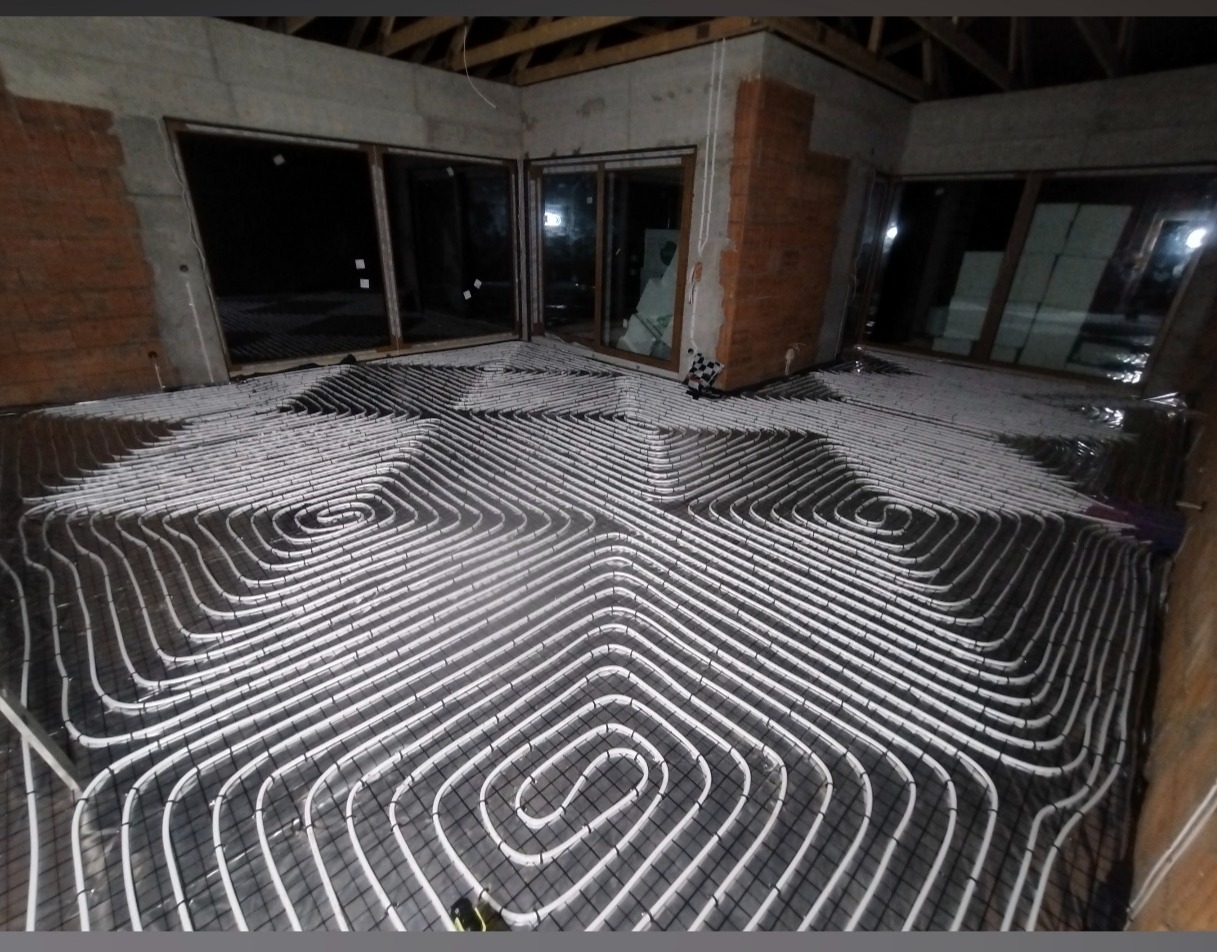 Białe rury ogrzewania podłogowego ułożone spiralnie na czarnej siatce w surowym wnętrzu z oknami. Widoczne elementy konstrukcyjne budynku.