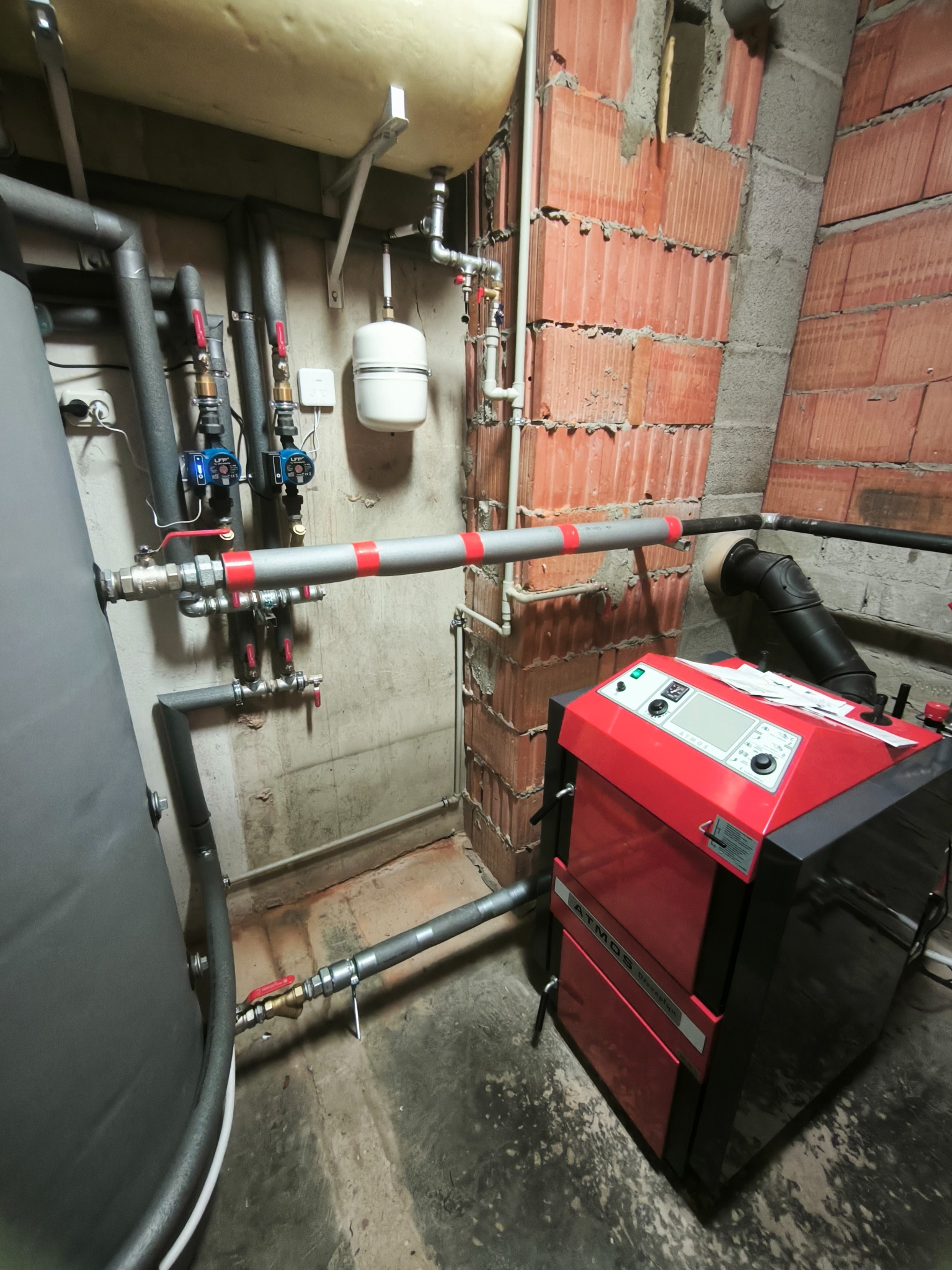 Czerwono-czarny piec CO marki Atmos z podłączonymi rurami instalacji grzewczej, widoczne elementy instalacji na ścianie z cegły i betonowej, szare rury z zaworami.