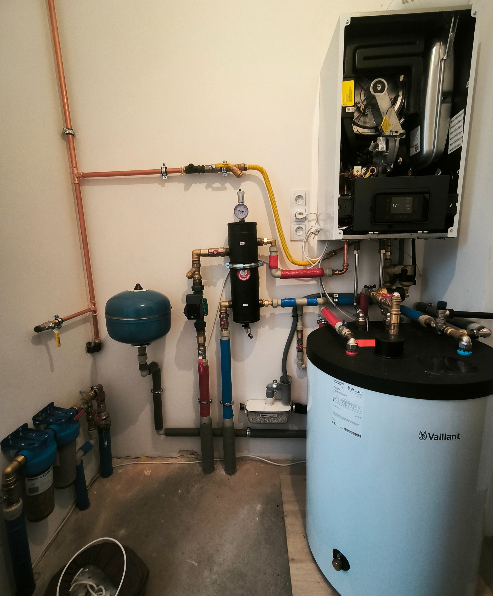 Złożona instalacja grzewcza z piecem gazowym Vaillant, zbiornikiem, filtrami i miedzianymi rurami w pomieszczeniu technicznym. Widoczne przyłącza i elementy sterujące.
