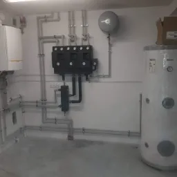 Kotłownia z kotłem gazowym, zasobnikiem ciepłej wody z kartonem na górze, licznymi rurami i armaturą w jasnym pomieszczeniu.