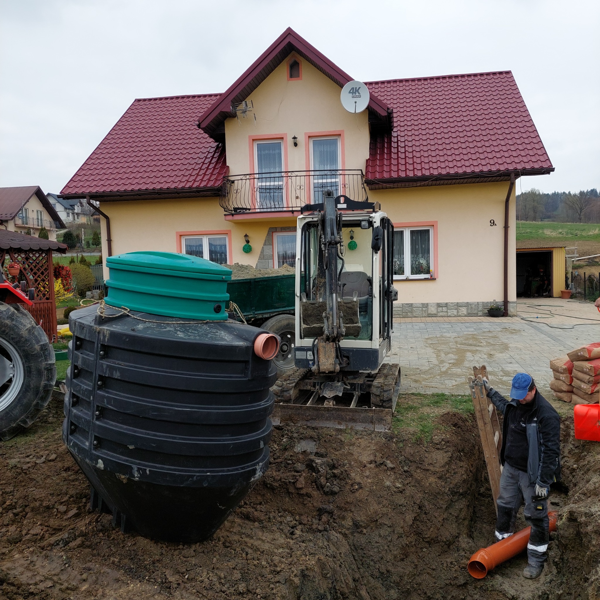 Montaż dużej, czarnej, plastikowej oczyszczalni ścieków z zieloną pokrywą obok wykopu, w tle dom z czerwoną dachówką i minikoparka, pracownik z rurą kanalizacyjną w wykopie.