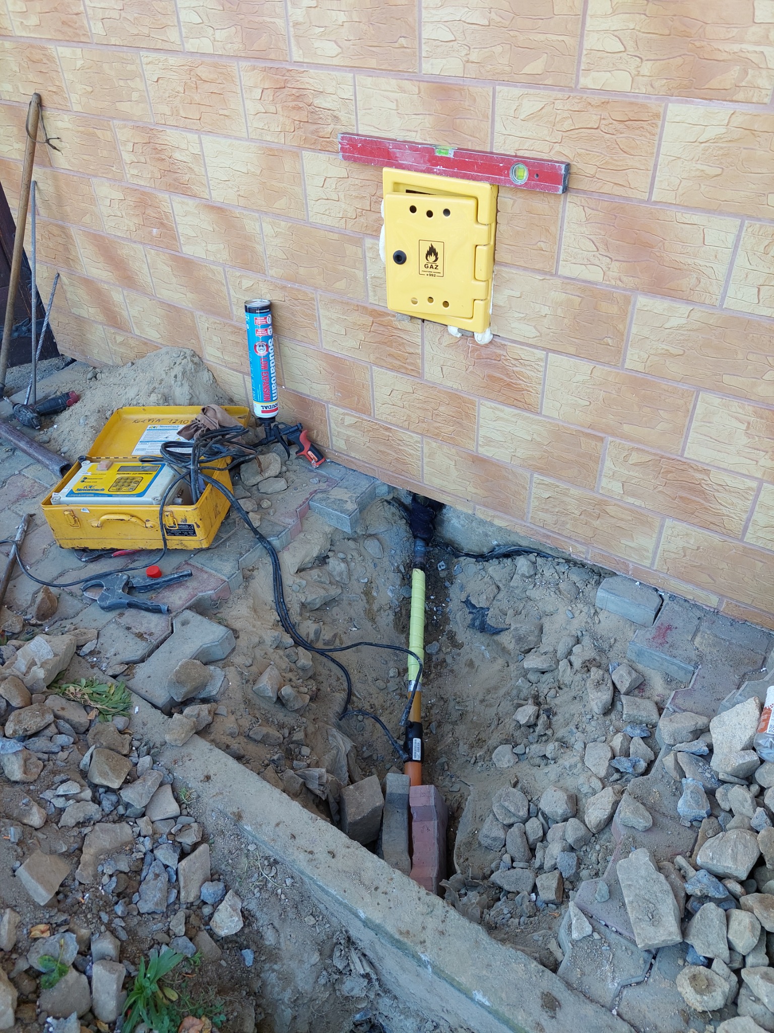 Żółta skrzynka gazowa z poziomnicą na elewacji budynku z imitacji cegły, widoczne wykopaliska i narzędzia podczas podłączania rury gazowej.