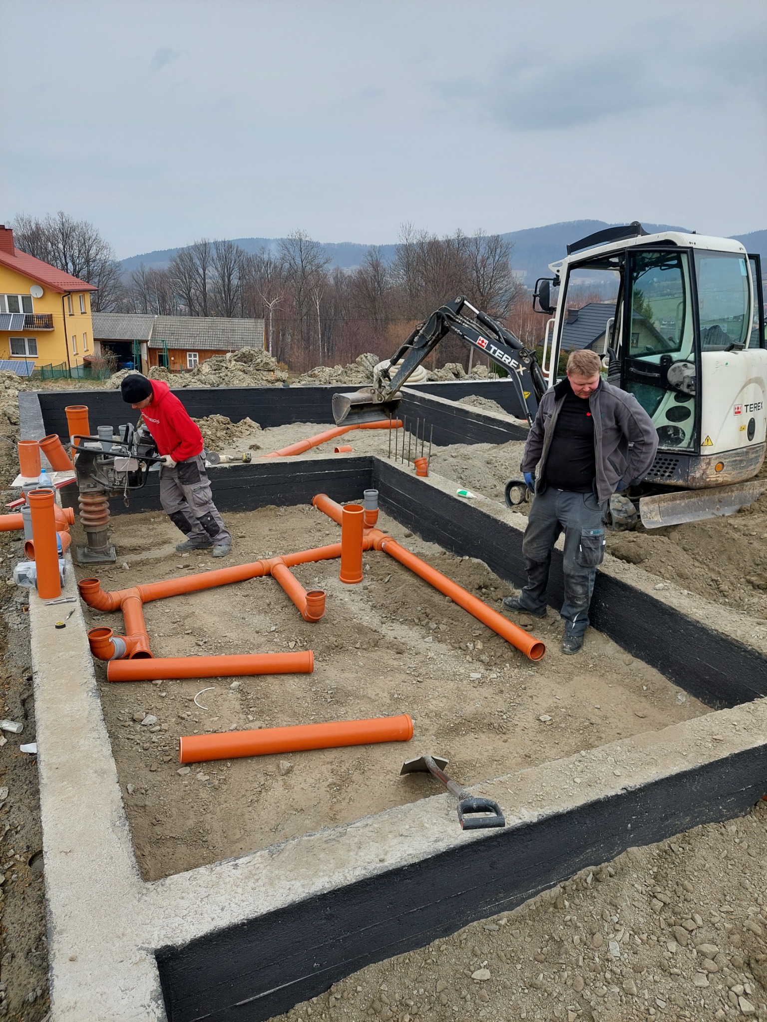Fundament budynku z hydroizolacją, wewnątrz rozłożone pomarańczowe rury kanalizacyjne, pracownik zagęszcza podłoże ubijarką, w tle minikoparka Terex i zabudowa wiejska.