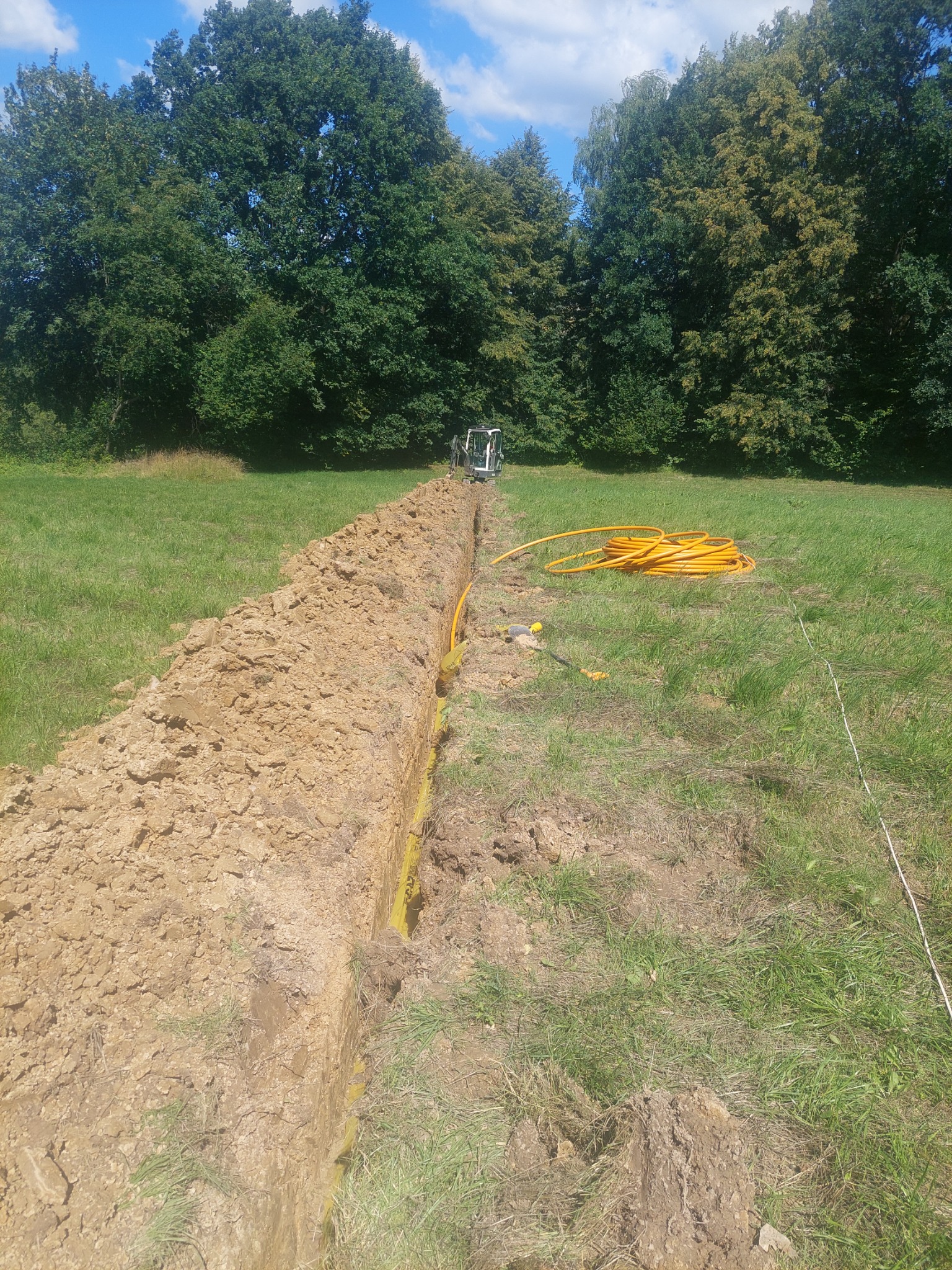 Wykopany rów w trawiastym terenie z ułożoną żółtą rurą, na tle zielonych drzew i błękitnego nieba z chmurami, z widocznym małym traktorkiem z tyłu.