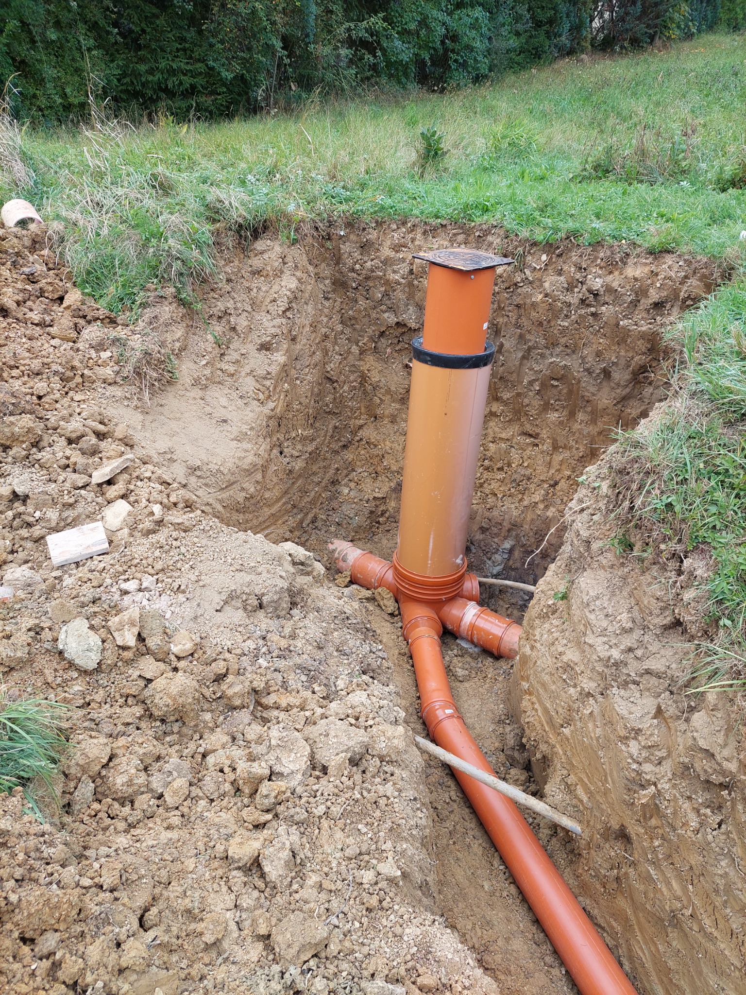 Pomarańczowe rury kanalizacyjne z tworzywa sztucznego ułożone w wykopie ziemnym na tle zielonej trawy, widoczne złącza i pionowy komin rewizyjny.