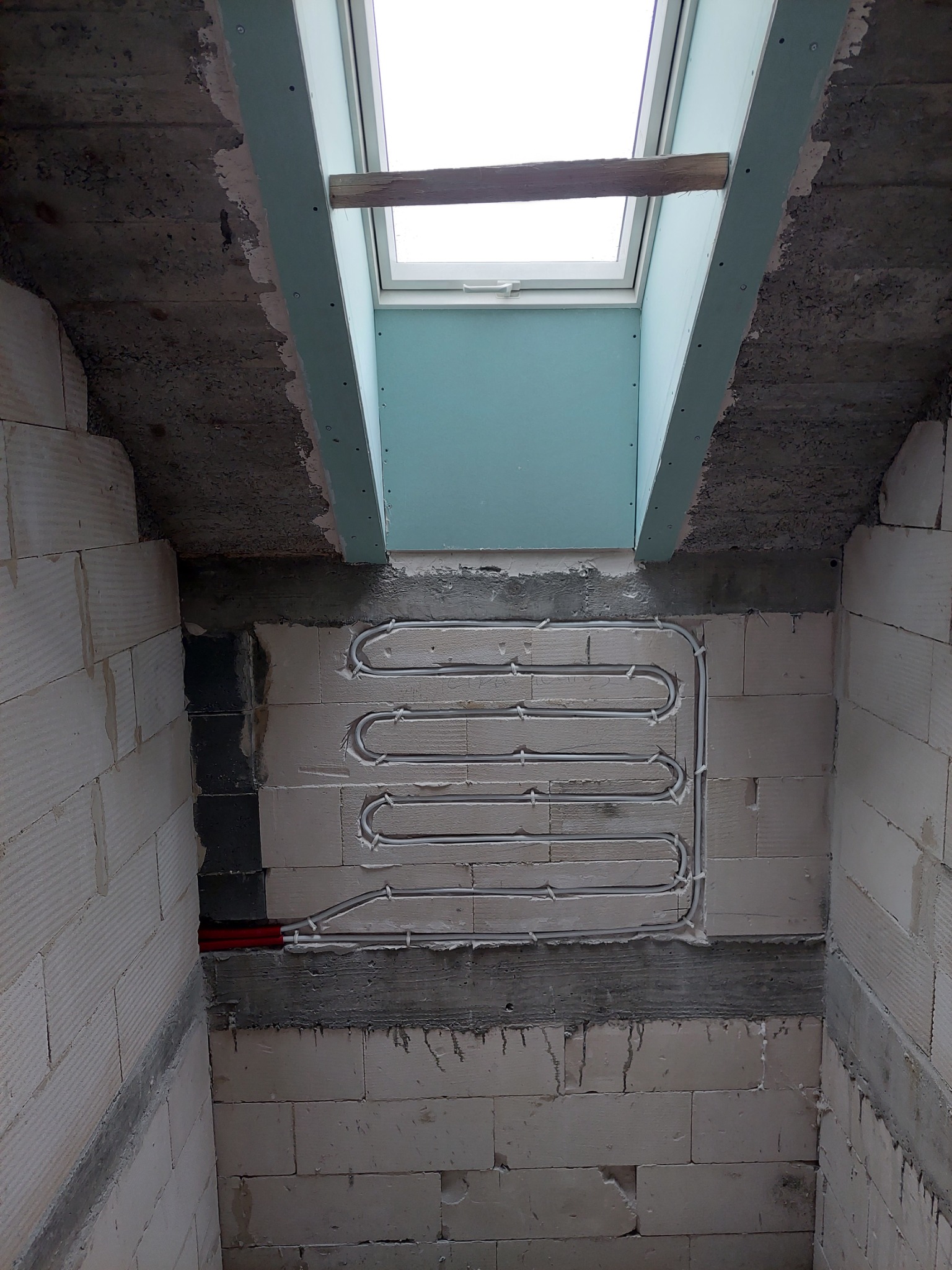 Montaż ogrzewania podłogowego w systemie wodnym, rurki ułożone w ścianie z bloczków betonu komórkowego, widoczne mocowania i fragment czerwonych rur doprowadzających, sufit z oknem dachowym w tle.