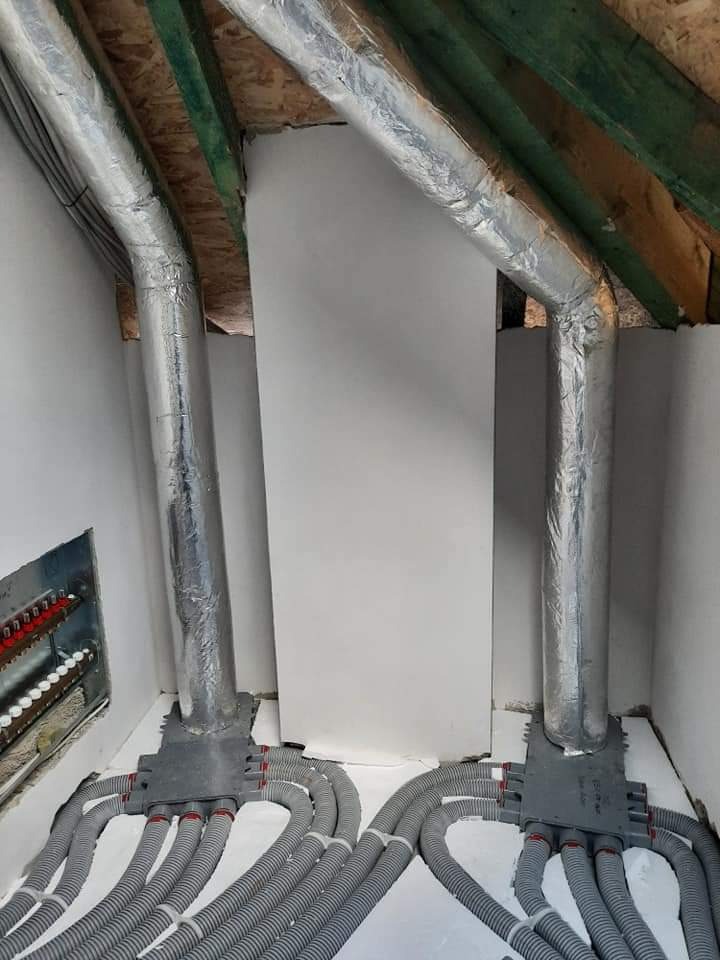 Dwie pionowe rury w srebrnej izolacji, połączone szarymi rozdzielaczami i licznymi elastycznymi przewodami, instalacja w surowym wnętrzu z widoczną drewnianą konstrukcją dachu.