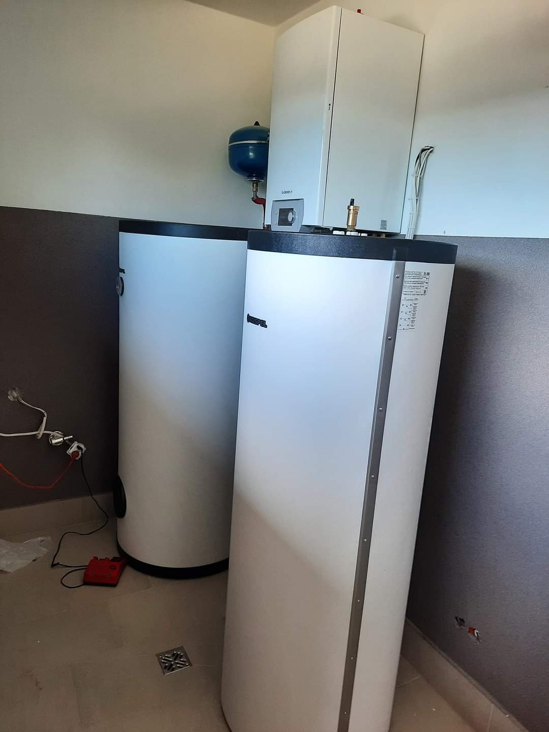 Dwa białe, cylindryczne zasobniki ciepłej wody w kotłowni, z widocznym niebieskim naczyniem wzbiorczym i fragmentem instalacji elektrycznej na ścianie.