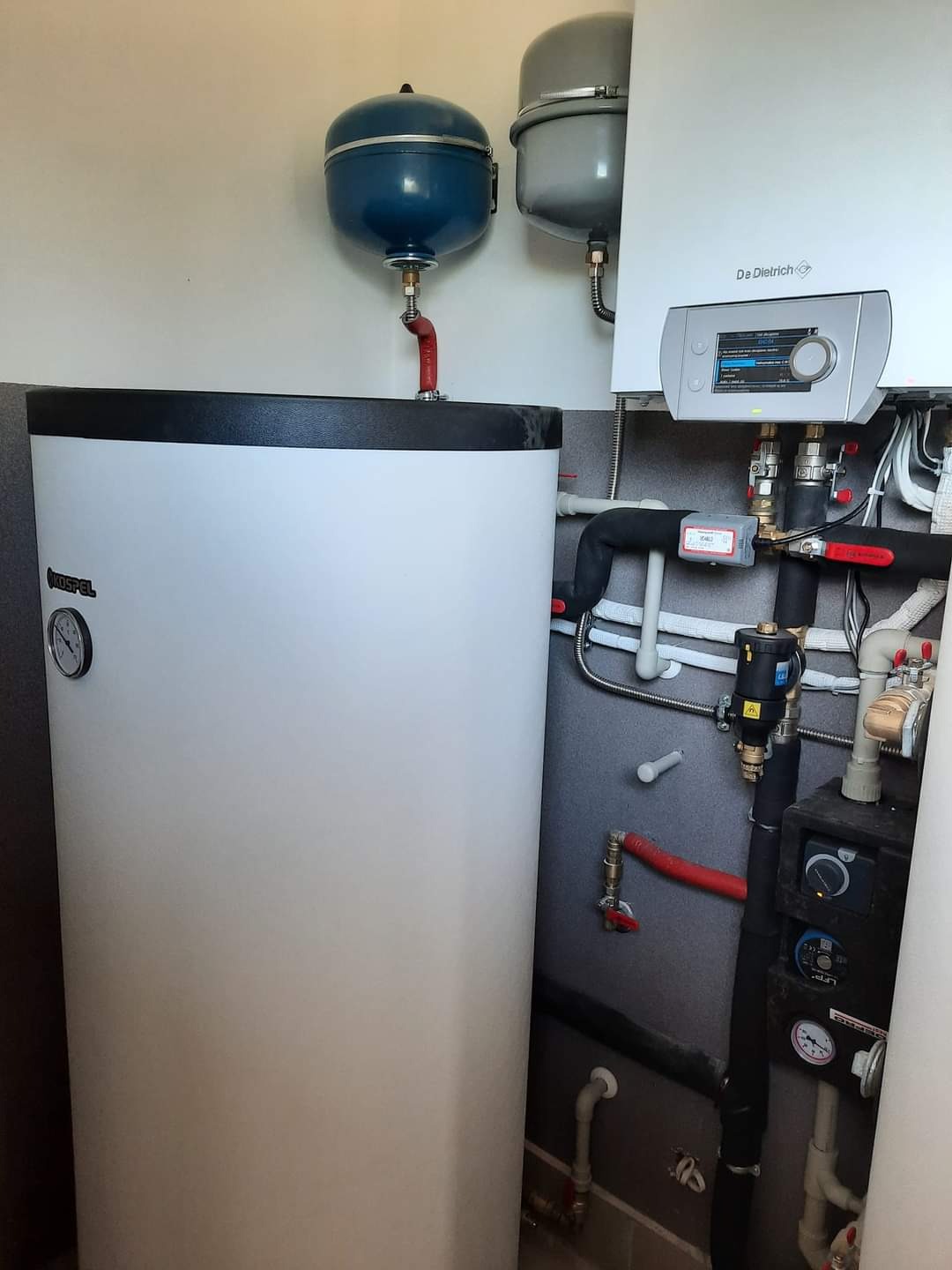 Kocioł grzewczy De Dietrich z podłączonym zasobnikiem Kospel, widoczne rury i elementy instalacji w pomieszczeniu technicznym.