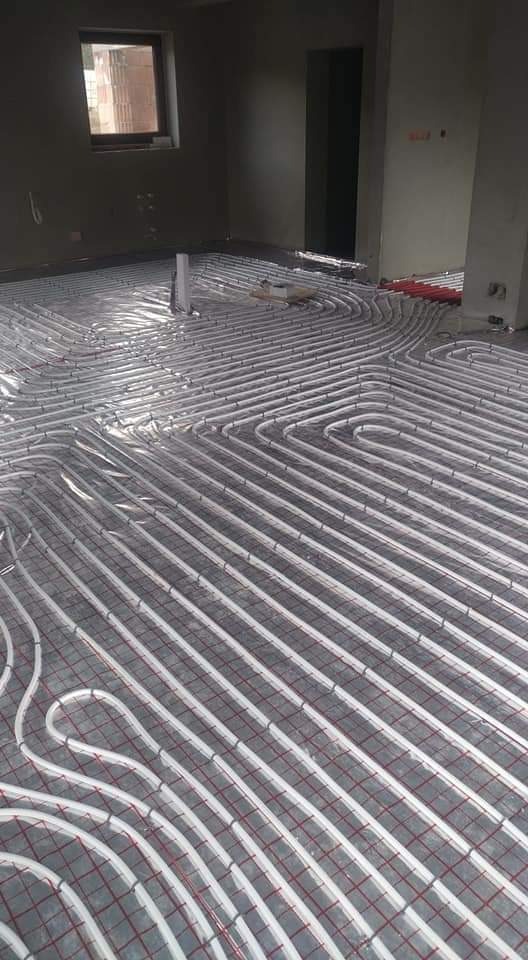 Białe rury ogrzewania podłogowego ułożone w pętle na siatce w surowym pomieszczeniu z oknem i szarymi ścianami, przygotowane do zalania wylewką.