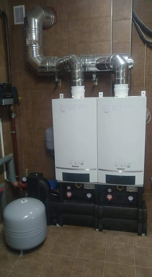 Dwa białe kotły grzewcze zamontowane obok siebie, z podłączonymi rurami odprowadzającymi spaliny, stojące na czarnej podstawie na tle brązowych płytek.