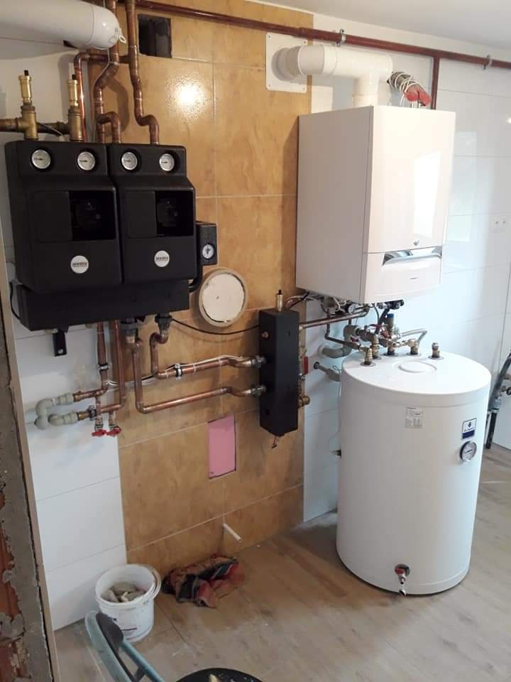 Kompletna instalacja grzewcza z dwoma czarnymi modułami sterującymi, białym piecem gazowym kondensacyjnym i białym zasobnikiem ciepłej wody, rury miedziane, brązowe i białe płytki ścienne, podłoga...