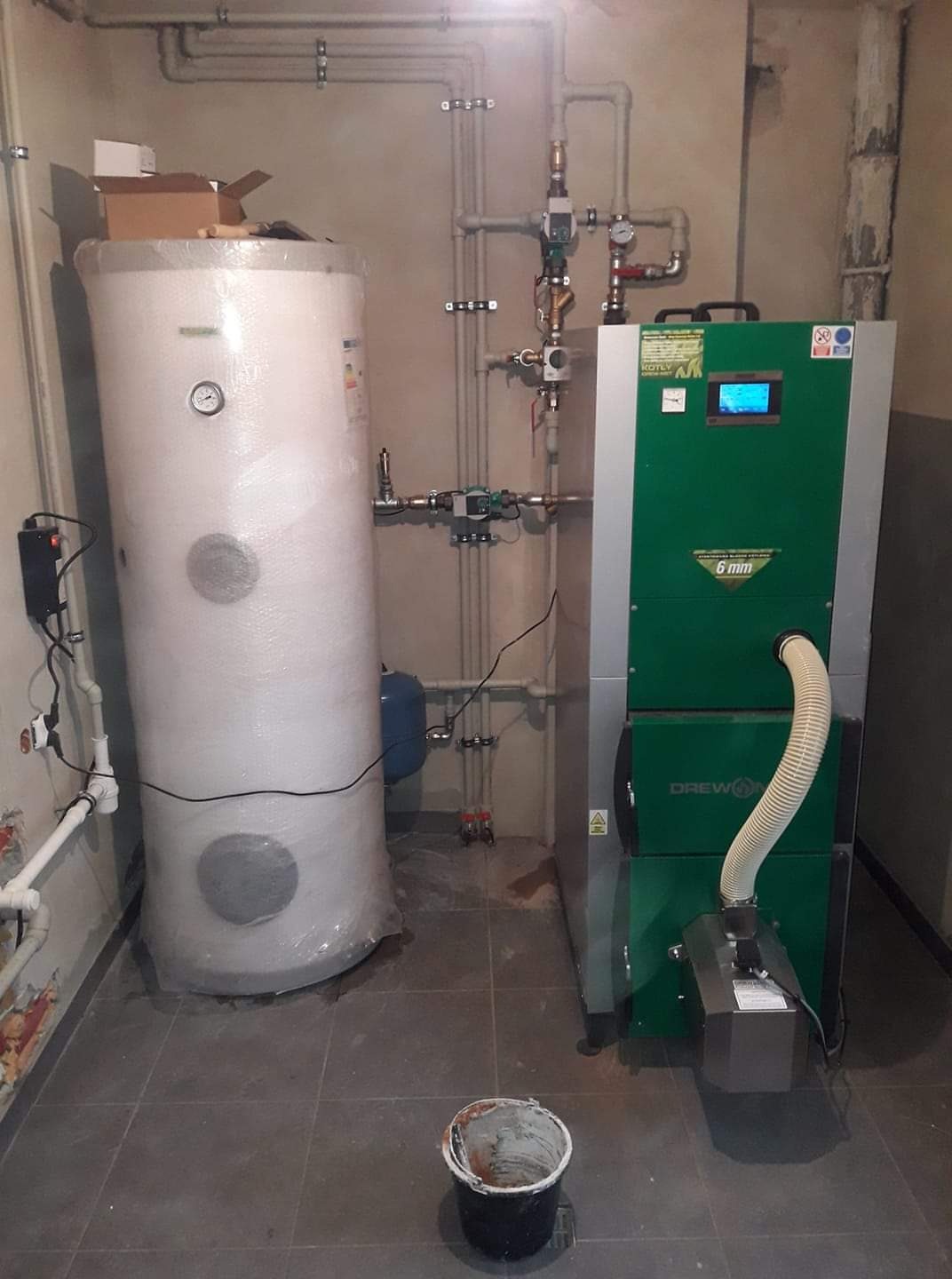 Nowoczesny, zielony piec Drewon z podajnikiem i zbiornikiem na wodę owiniętym folią, widoczne rury instalacji grzewczej i wiadro na podłodze w pomieszczeniu technicznym.