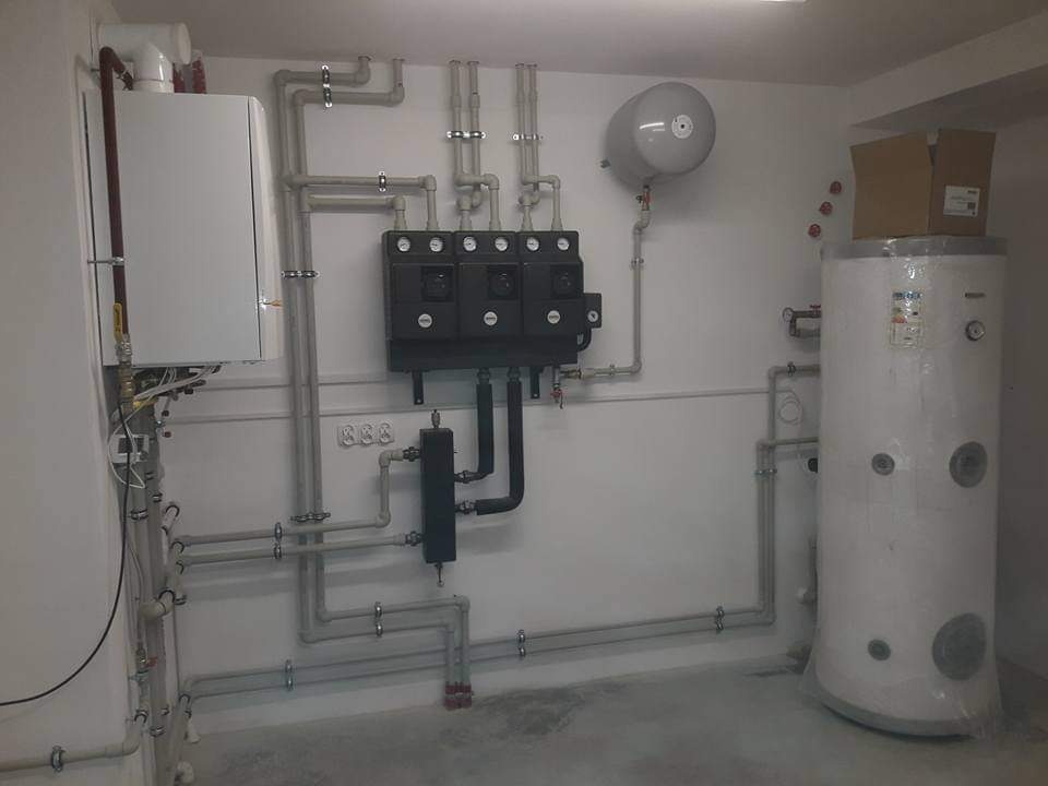 Kotłownia z białym kotłem, szarymi rurami, czarną rozdzielnicą i dużym białym zbiornikiem na wodę, częściowo owiniętym folią ochronną, w jasnym pomieszczeniu.