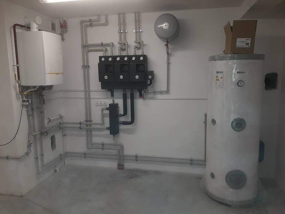 Kotłownia z kotłem gazowym, zasobnikiem ciepłej wody z kartonem na górze, licznymi rurami i armaturą w jasnym pomieszczeniu.