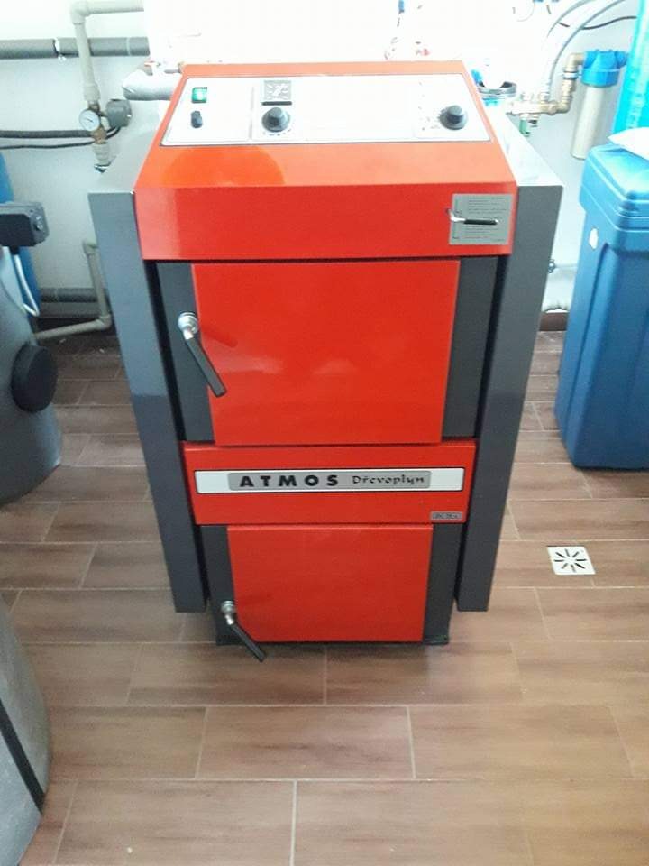 Czerwono-szary piec Atmos Drevo plyn na drewno, z widocznymi rurami instalacji w tle, zainstalowany na brązowych płytkach podłogowych.