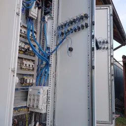 Otwarta szafa sterownicza z widocznymi elementami elektrycznymi, licznymi przewodami w kolorze niebieskim i żółtym, zamontowana na zewnątrz budynku, z widocznym fragmentem dachu i nieba w tle.