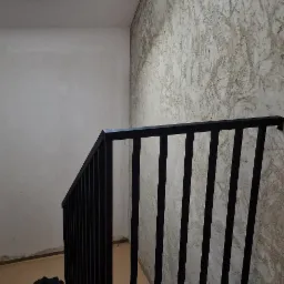 Nowoczesna, czarna balustrada schodowa z pionowymi szczeblami. Widoczne schody z drewnopodobnych paneli i dekoracyjna faktura ściany. Ujęcie z dołu, z oknem dachowym.