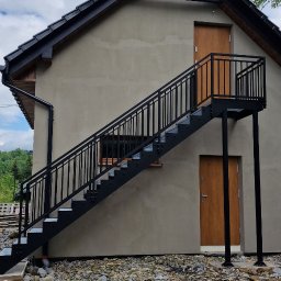 Siepak - Stalowe Realizacje - Zewnętrzne schody metalowe z czarną balustradą prowadzące do drzwi na piętrze budynku z szarą elewacją. Widoczne kamienie na podłożu i fragmenty zieleni w tle.