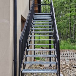 Siepak - Stalowe Realizacje - Zewnętrzne schody metalowe z czarną poręczą, prowadzące na piętro budynku w Budzowie. Widok z dołu, na tle zieleni.