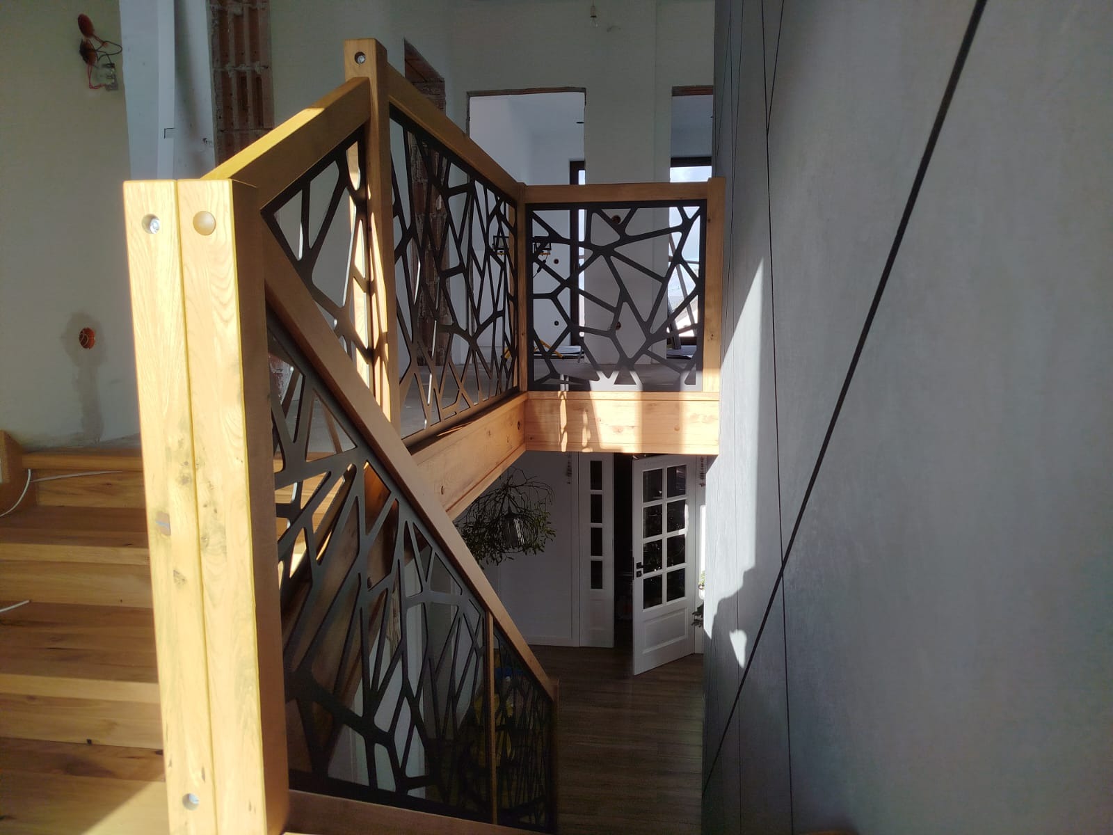 Drewniane schody z nowoczesną, czarną balustradą o ażurowym wzorze, prowadzące na dół. Widok z góry, jasne oświetlenie, minimalistyczny design.