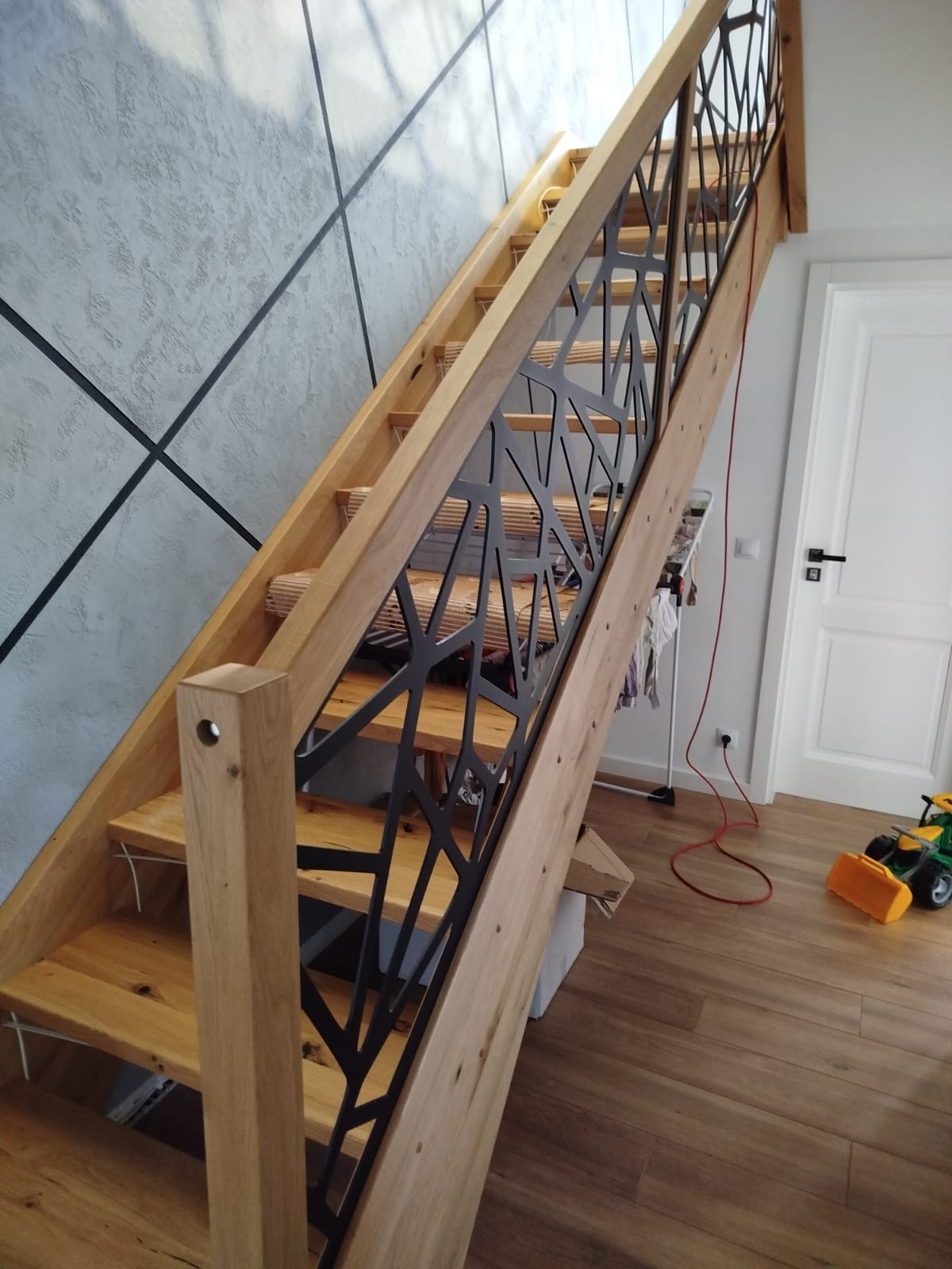 Drewniane schody z oryginalną, metalową balustradą o geometrycznym wzorze, w nowoczesnym wnętrzu z akcentem na betonową ścianę i drewnianą podłogę.