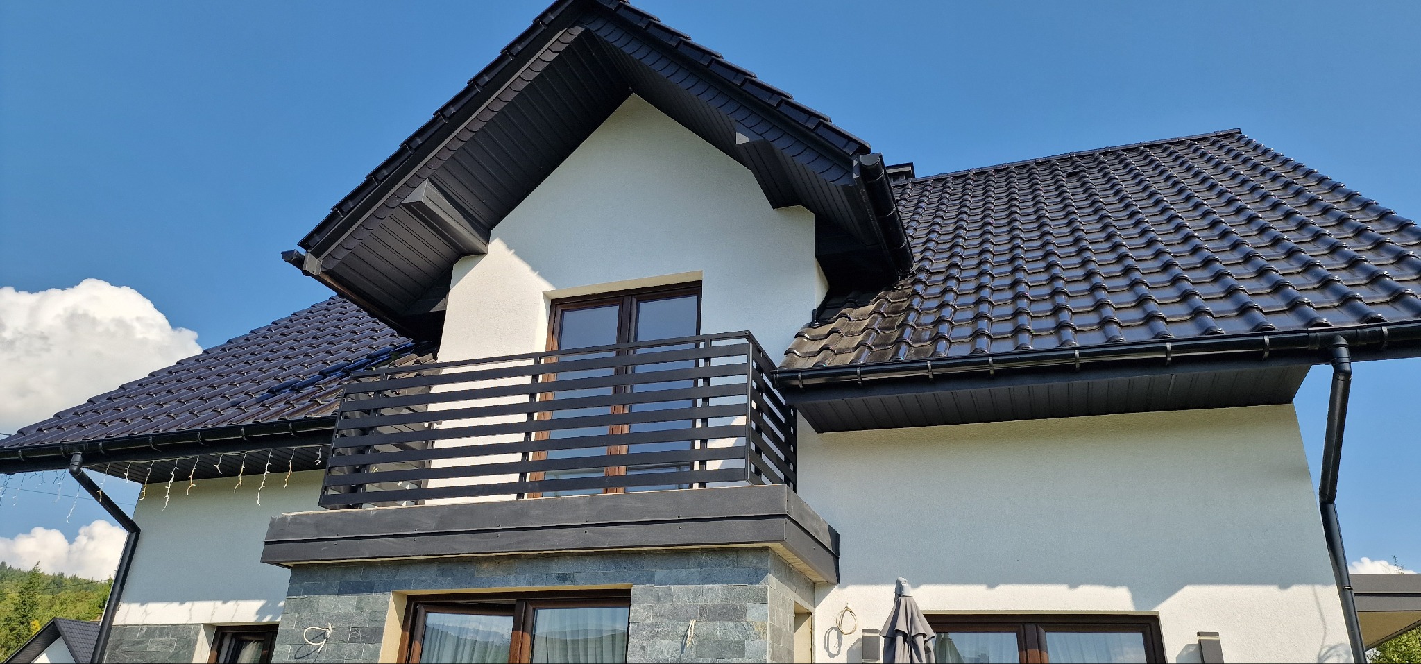 Nowoczesny dom z balkonem i czarną balustradą. Widoczna dachówka w tym samym kolorze i jasna elewacja. Ujęcie z dołu, błękitne niebo w tle.