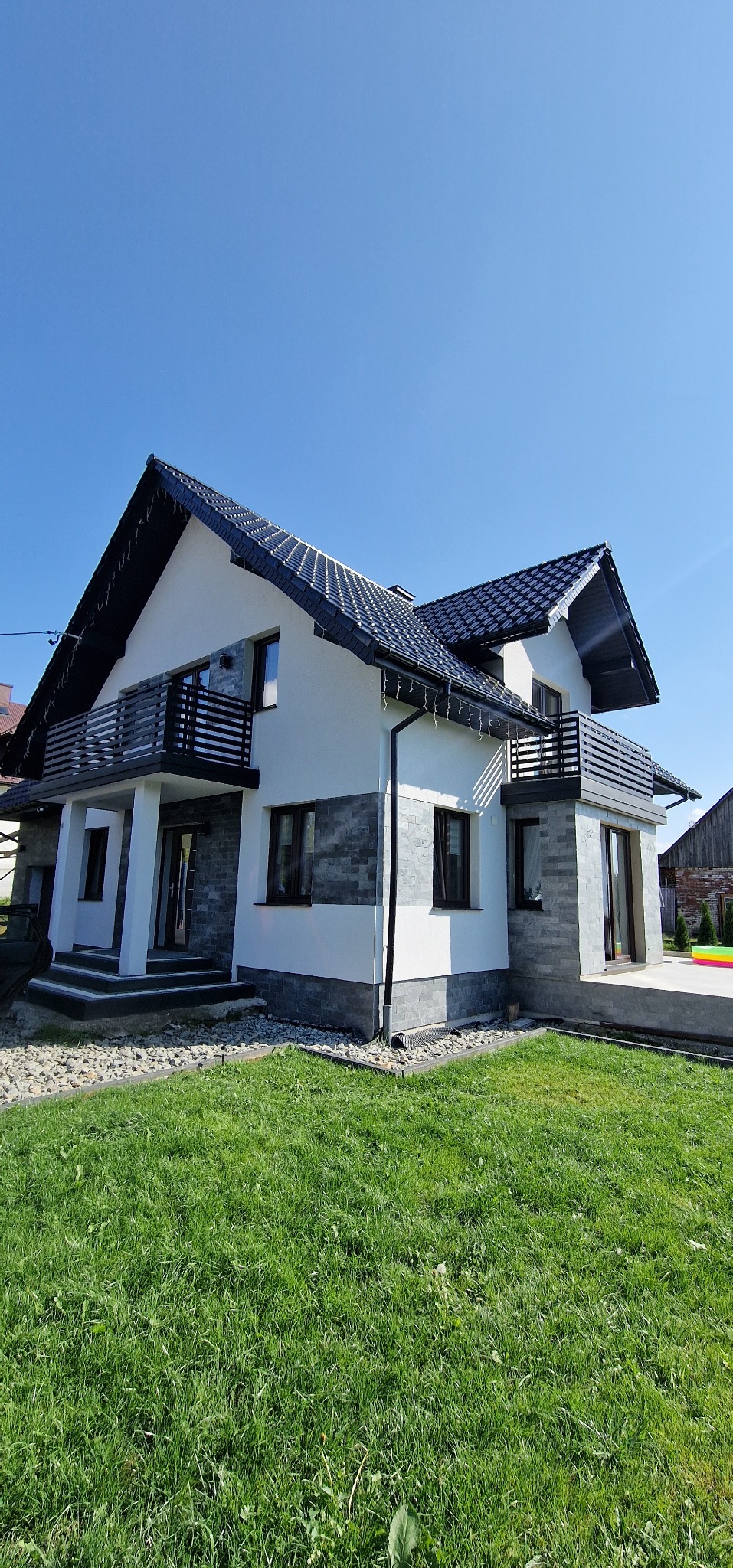 Dwukondygnacyjny dom z balkonami z czarnymi, poziomymi balustradami. Elewacja w odcieniach bieli i szarości, zielony trawnik przed budynkiem i błękitne niebo.