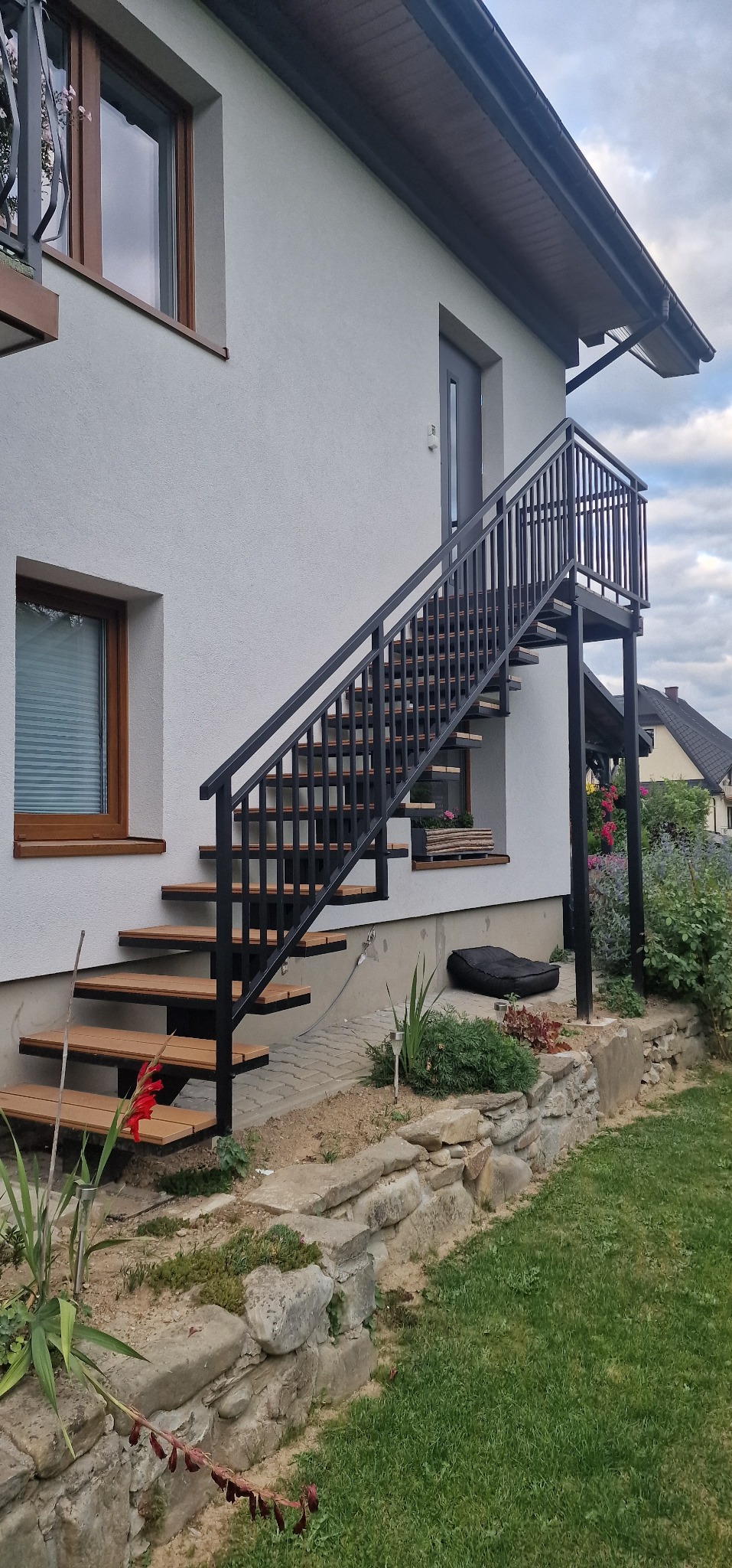 Zewnętrzne schody z metalową konstrukcją w kolorze czarnym i drewnianymi stopniami, prowadzące do drzwi na piętrze, z widoczną roślinnością w otoczeniu.