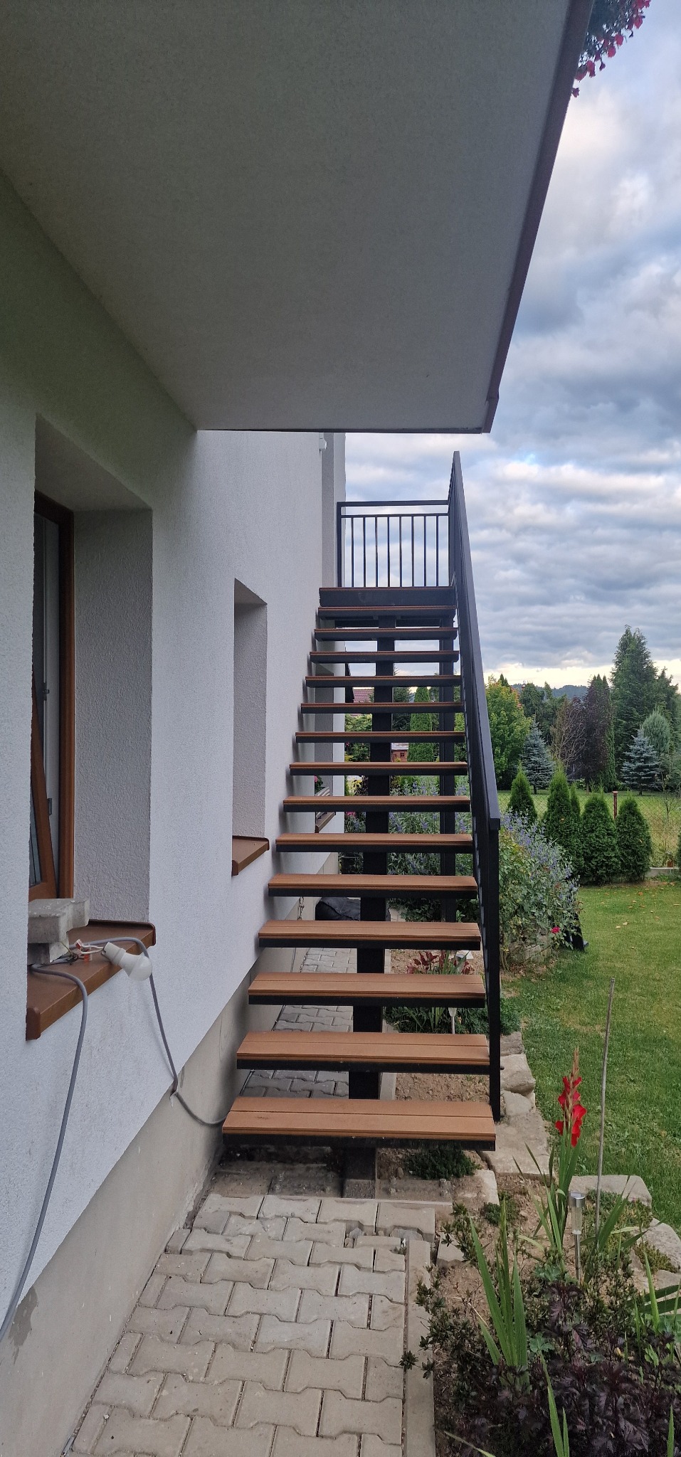 Zewnętrzne schody metalowe z drewnopodobnymi stopniami, prowadzące do wejścia domu. Czarne poręcze i balustrada. Widok na ogród i pochmurne niebo.