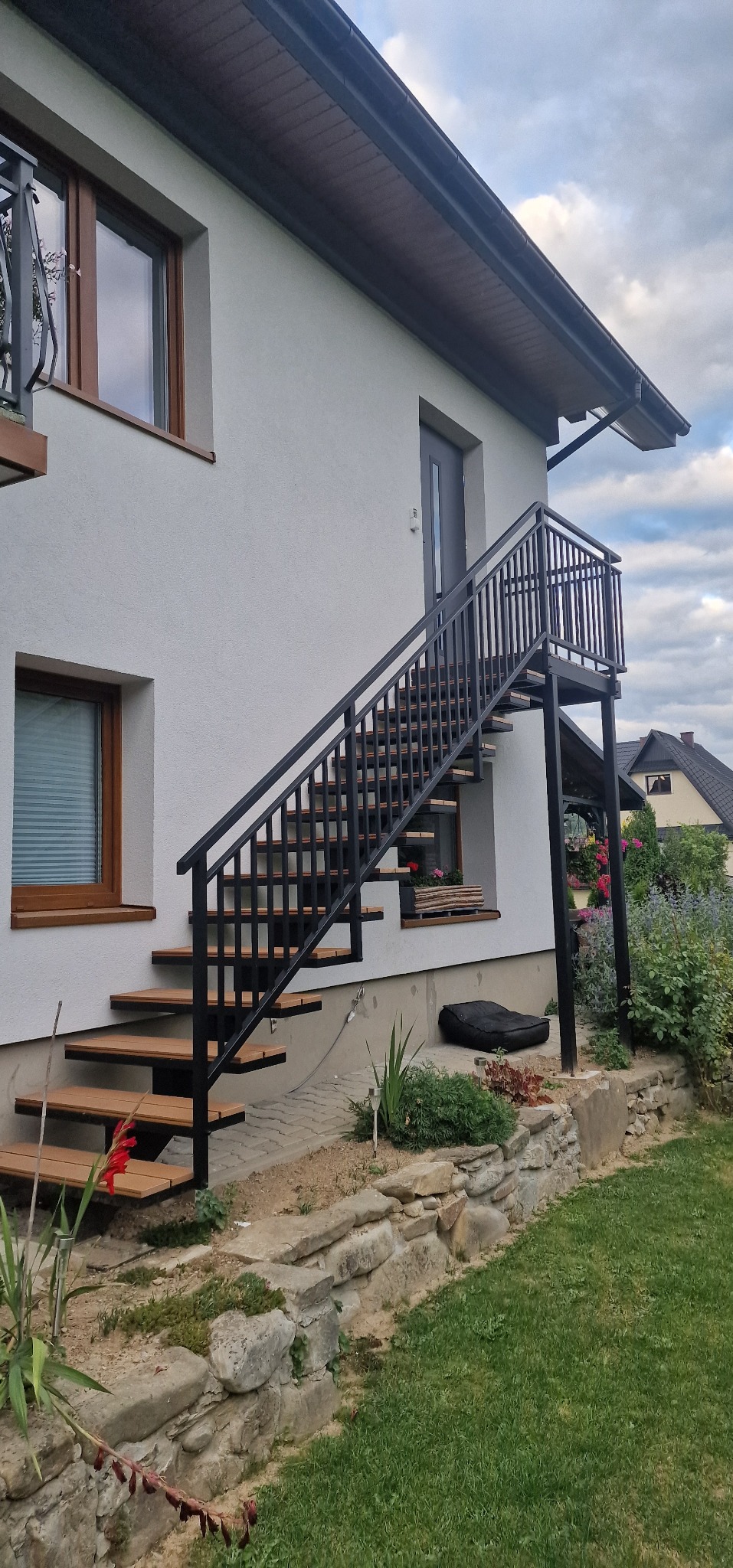 Zewnętrzne schody z czarną metalową konstrukcją i drewnianymi stopniami, prowadzące do drzwi na piętrze. Widok na dom z ogrodem.