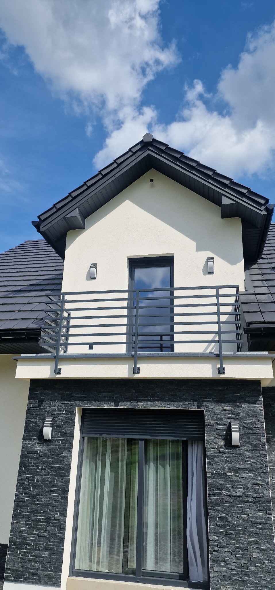 Nowoczesna balustrada balkonowa z poziomych profili na tle elewacji domu z oknem i fragmentem dachu pod błękitnym niebem.