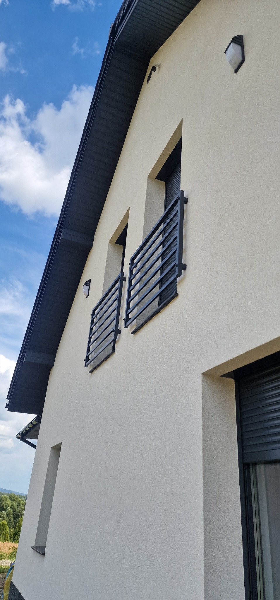 Elewacja domu z dwoma czarnymi, metalowymi balustradami balkonowymi. Widok z dołu, błękitne niebo z chmurami w tle. Minimalistyczny design.
