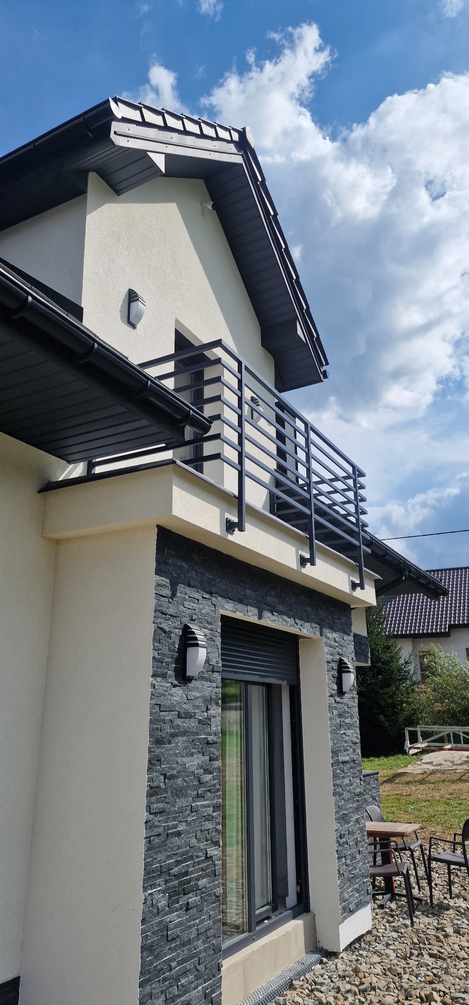 Nowoczesny dom z balkonem i czarną, prostą balustradą. Elewacja z kamienia dekoracyjnego i oświetlenie zewnętrzne. Ujęcie z dołu, widok na błękitne niebo.