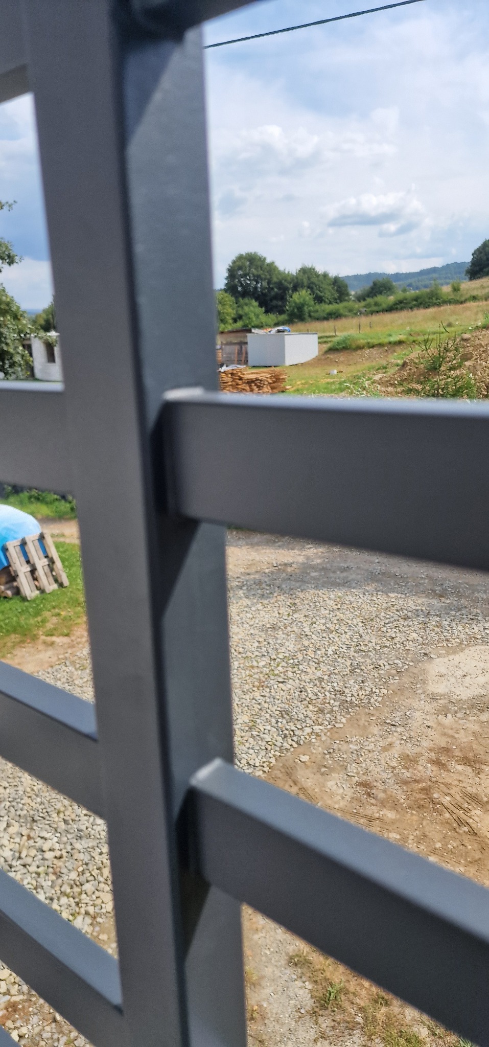 Metalowa konstrukcja z widokiem na wiejski krajobraz z budynkami gospodarczymi i stertą drewna w tle, uchwycona w słoneczny dzień.