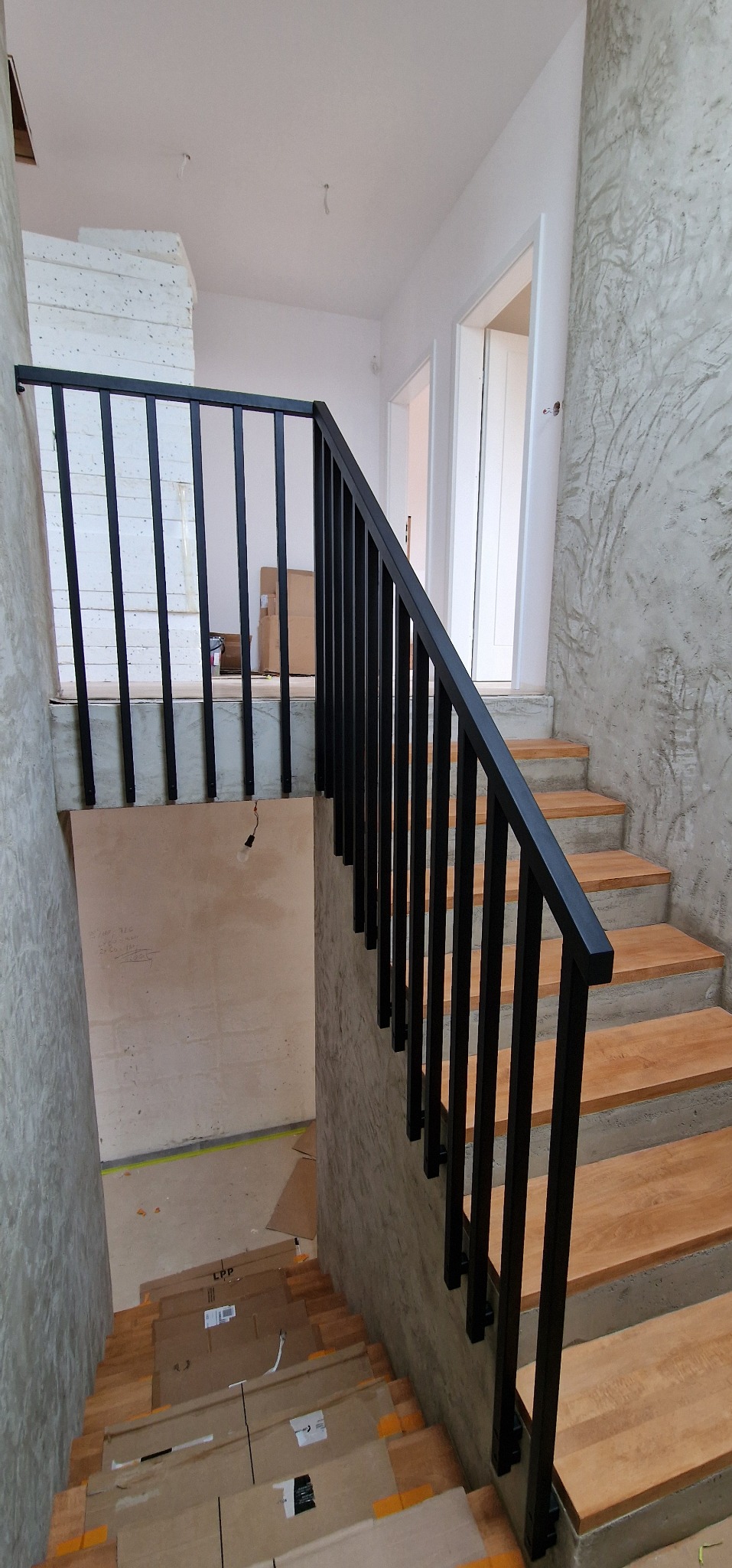 Nowoczesne schody z drewnianymi stopniami i czarną, minimalistyczną balustradą. Widoczne surowe ściany i zabezpieczenia na stopniach.